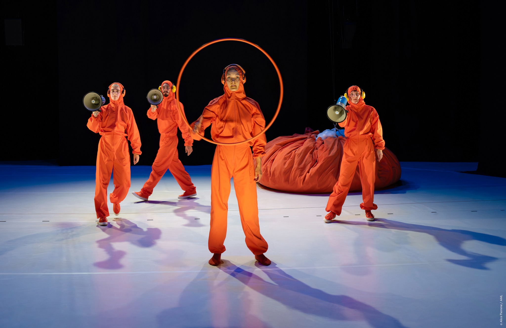 4 people on stage with orange overalls, person in the middle holding a hula hoop